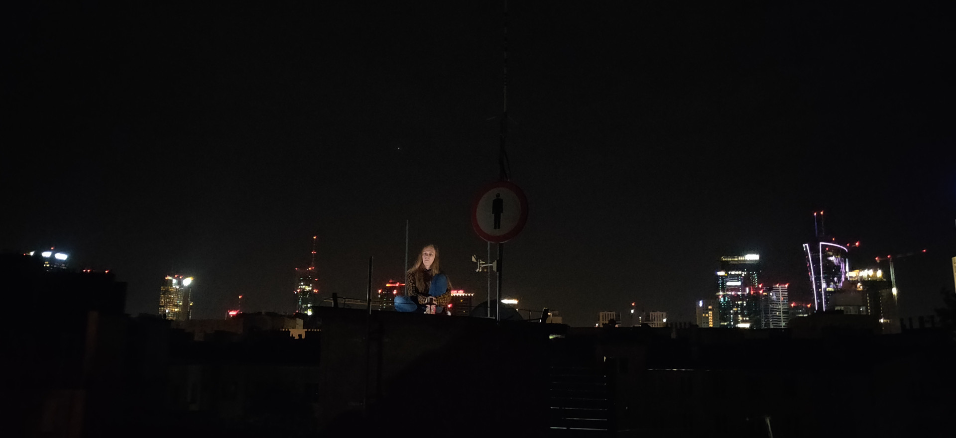 A photo of me sitting on the roof of a building in Warsaw - specifically, the one of the Warsaw Hackerspace. Behind me, you can see the skyline of Warsaw's Wola district. Photo credits: @erin@quiescent.nexus on the Fediverse.